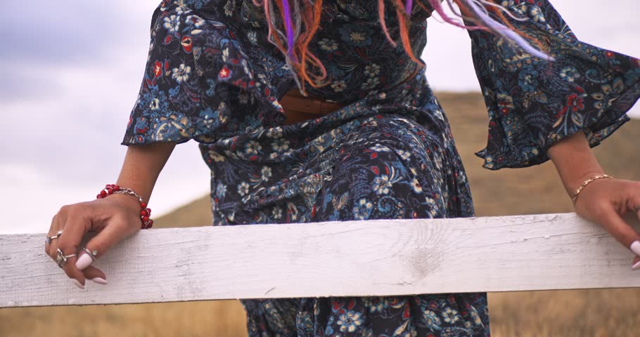 A woman in boho style with dreadlocks leans her hands on a rustic fence. Close-up on her hands