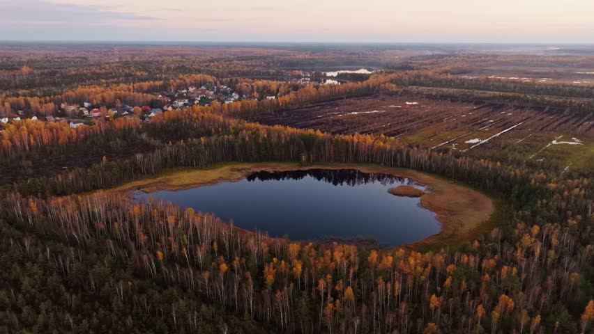 Aerial view of calm bog lake and village at sunset, surrounded by autumn forest.