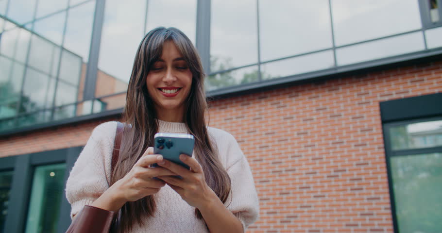 Camera front view of happy Caucasian girl walking away from modern college. Student walking back home. Using her smartphone with both hands. Texting. Typing message on touch screen.