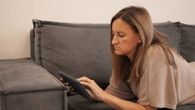 Woman lying on couch browsing on digital tablet, fully engaged in content. Digital tablet offers convenience and easy access to media. Ideal for themes of relaxation, tech, and digital tablet usage. - Powered by Shutterstock - Get 15% off with code: PIKWIZARD15