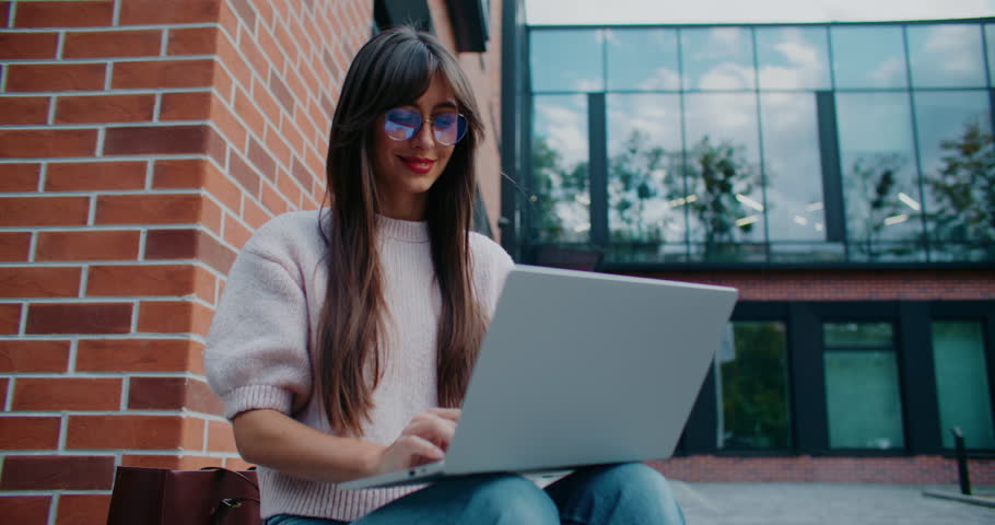 Camera moving closer to Caucasian girls face. Woman working remotely on her laptop. Typing on keyboard. Screen of portative computer mirroring from woman's glasses. Modern building with tall windows.