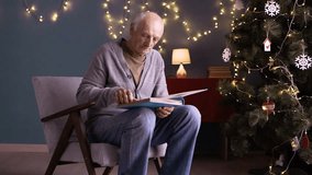 Elderly man sitting in cozy chair, looking through old family photo album during Christmas time. Christmas tree with decorations and warm lights in background - Powered by Shutterstock - Get 15% off with code: PIKWIZARD15