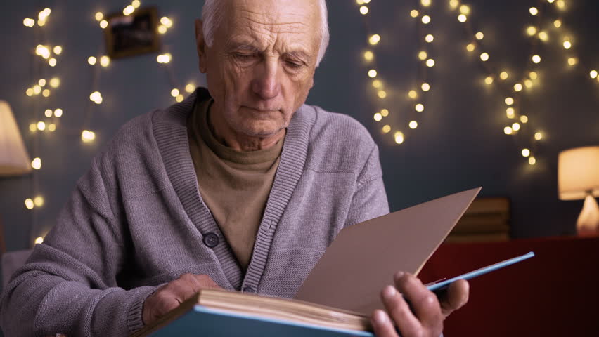 Retired man looking at old photographs in photo album, reflecting on memories from past. Surrounded by warm holiday lights, he appears deep in thought, reminiscing with nostalgia.