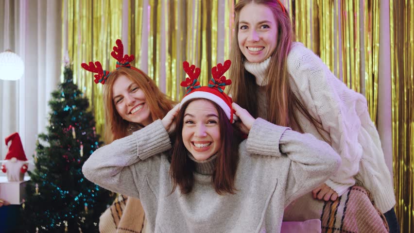 Three friends enjoy holiday fun in a cozy room. Two women place a Santa hat on their friend, sharing laughs and hugs amid festive Christmas decorations and twinkling lights