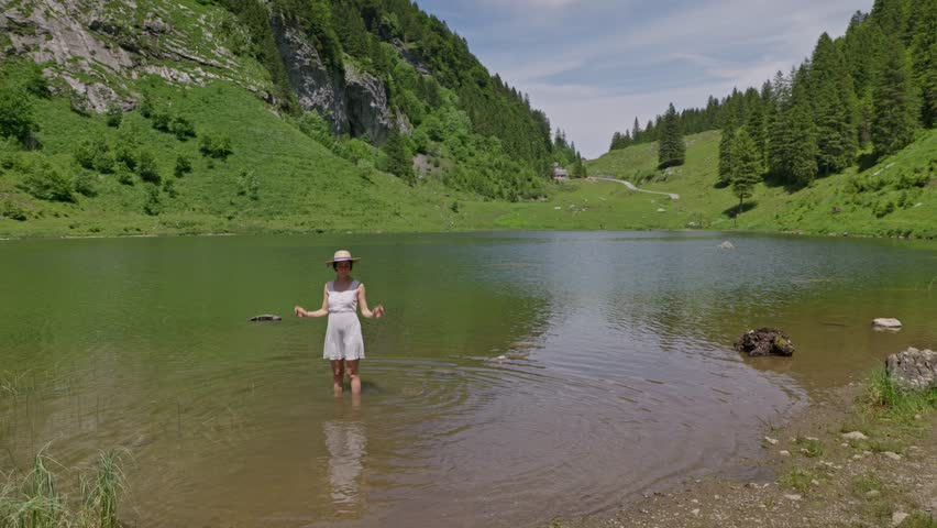 A woman in a white dress wades in Talalpsee Lake, surrounded by lush greenery and mountains