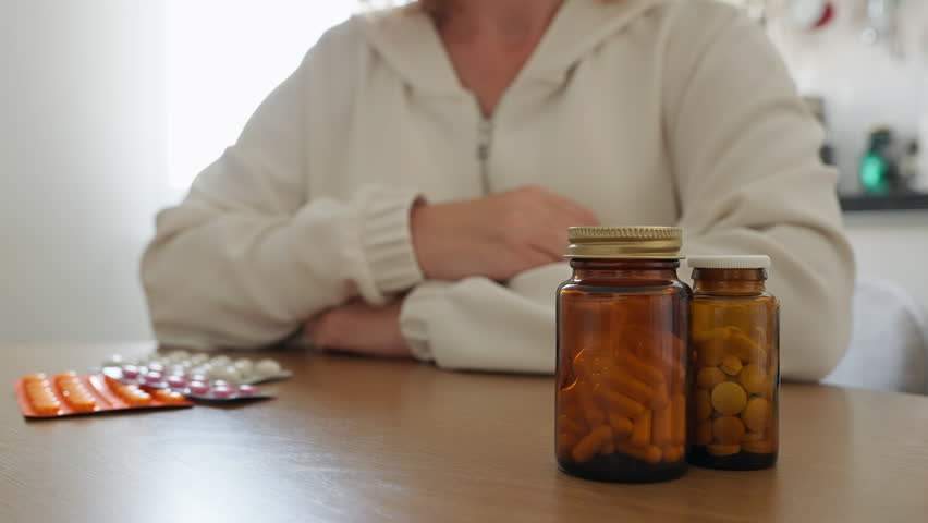 Female pours supplement capsules from brown glass bottle into their hand, surrounded by other bottles and blister packs on table. Self-care, health, daily wellness routines. Women's health. Lifestyle 