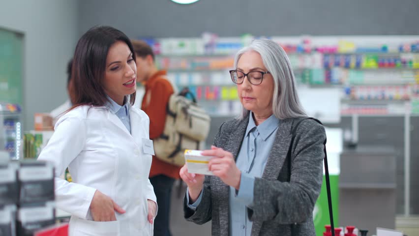 Female pharmacist advising medicine to female client. Woman druggist advising pills to older customer. Caucasian pharmacist and customer in pharmacy. Senior woman choosing drugs in drugstore.