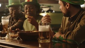 Side chest up shot of group of three diverse friends in green hats sitting at bar counter with snacks and ale, talking and hanging out in evening in pub on St Patricks day - Powered by Shutterstock - Get 15% off with code: PIKWIZARD15