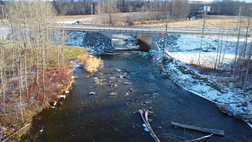 A serene winter landscape with a river flowing under a road bridge, surrounded by snow-covered banks and bare trees. Sayabec, Quebec, Canada, 2024.