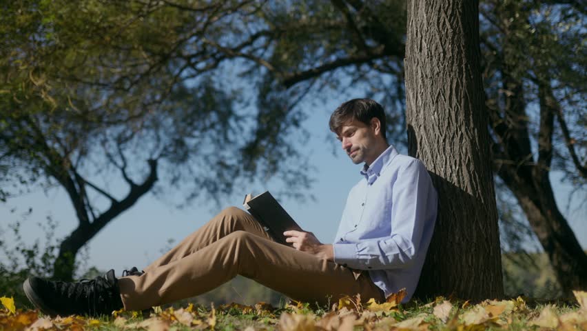 Middle-aged man reflects by the lakeside, seated on autumn leaves, with a book in hand, embracing the calm atmosphere of fall in an outdoor setting.
