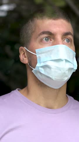 Caucasian young adult man wearing facemask behind glass window at home. Quarantine concept. Vertical video