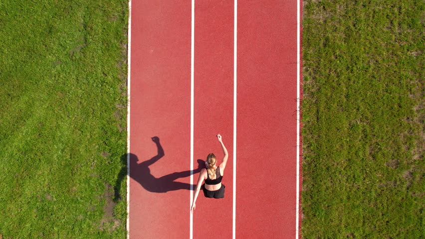 A single runner on a red track surrounded by green grass, captured from above. The strong shadow emphasizes the movement