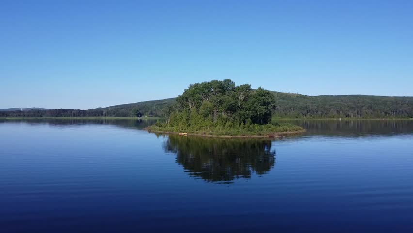 Serene image of a lush green island in the middle of a peaceful lake, reflecting clear blue skies and distant hills. Ideal for concepts of tranquility, nature, and solitude.