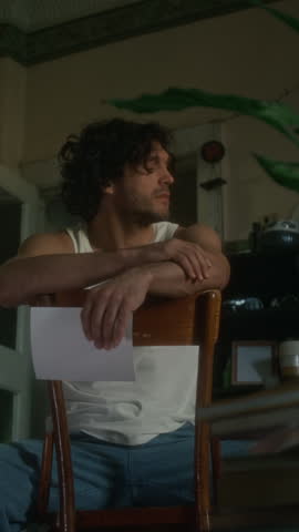 Vertical shot of depressed and tired Caucasian man in undershirt and jeans sitting on chair with photograph and thinking about meaninglessness of life, feeling lonely, spending days in shabby flat