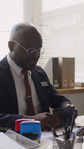 Vertical shot of confident Black male visa officer in suit and glasses interviewing offscreen applicant in consulate