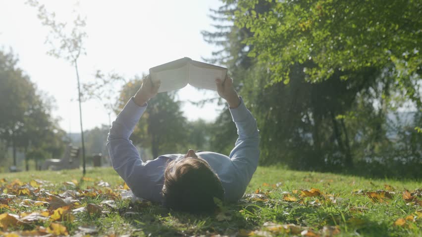 In a serene park setting, a man lies back on the grass with a book in hand, enjoying the gentle warmth of autumn. Surrounded by trees and fallen leaves, he finds peace in this quiet moment outdoors.