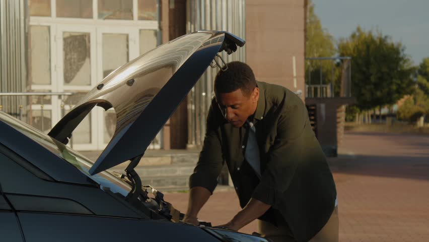 Handsome African American male driver in casual clothes standing near open car hood, trying to fix broken vehicle engine while driving