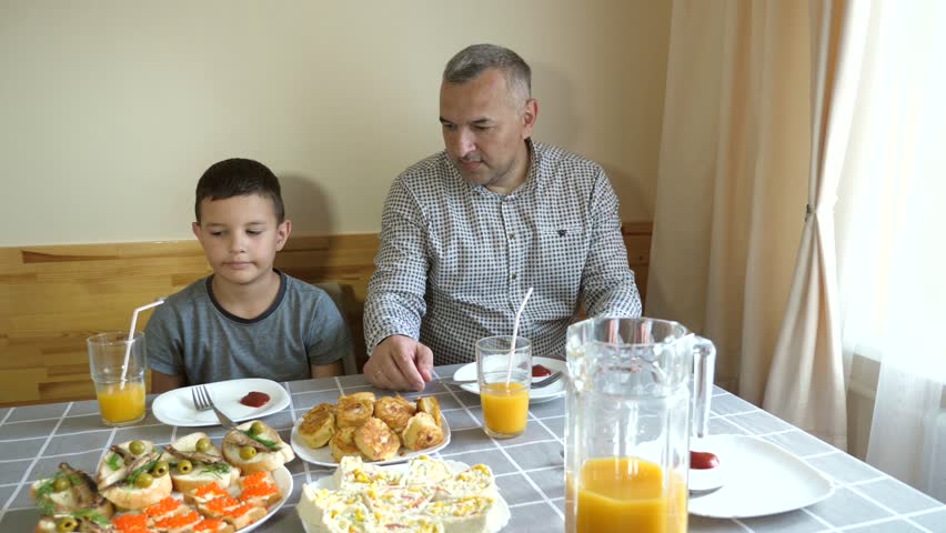 Father and son are sitting at the table, eating food and drinking drinks.