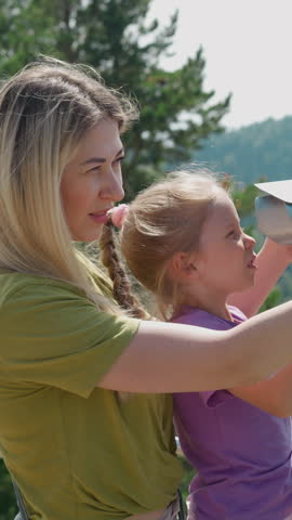 Long haired blonde woman adjusts tower viewer and lifts up little daughter to look through tool on ground at highland resort slow motion