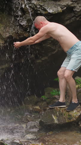Joyful man tourist wets bald head with clear waterfall streams standing on old slippy rock at virgin highland reserve on summer day slow motion