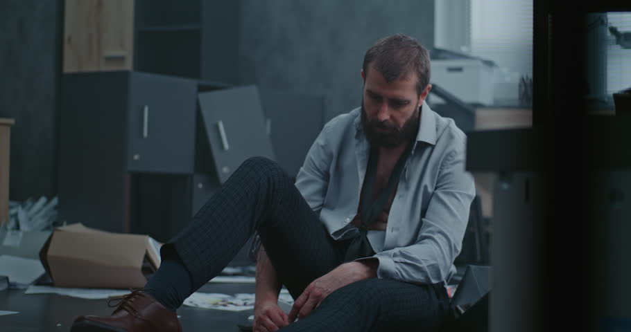 Frustrated office worker sits on the floor, breathes heavily after destroying office room. Depressed businessman after fit of rage over unsuccessful business deal. Financial collapse. Job burnout.