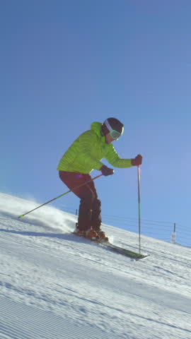 TRACKING SLOW MOTION: Young adult happy skier doing fast turns on a fresh winter morning groomed piste at European ski resort in the Alps during cloudless day. Sun flare shinning in the camera.
