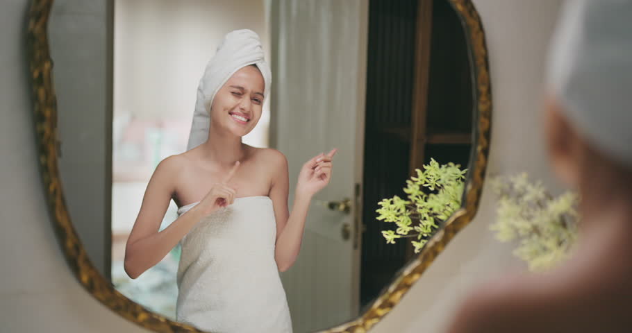 Indian young adult gen z smiling beautiful woman wrapped towel on head and body look bathroom mirror speak to her self reflection dancing sing song enjoy morning good mood having fun joy indoor home