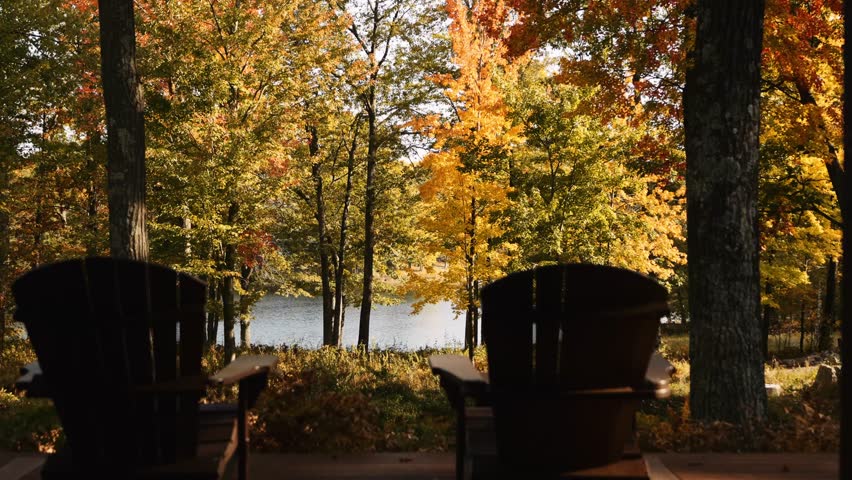 A scenic view of two Adirondack chairs overlooking a lake framed by vibrant autumn trees with golden and green leaves