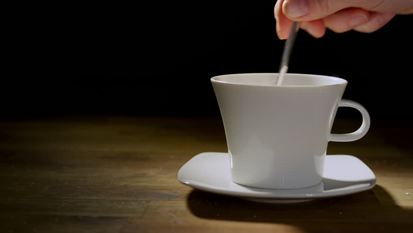 Male hand adding sugar into the white cup using a teaspoon, on the black backdrop, close up shot. Bad habit, food and drink concepts.