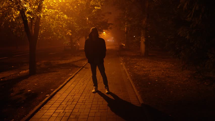 Mysterious figure stands on a foggy street at night, illuminated by street lamps