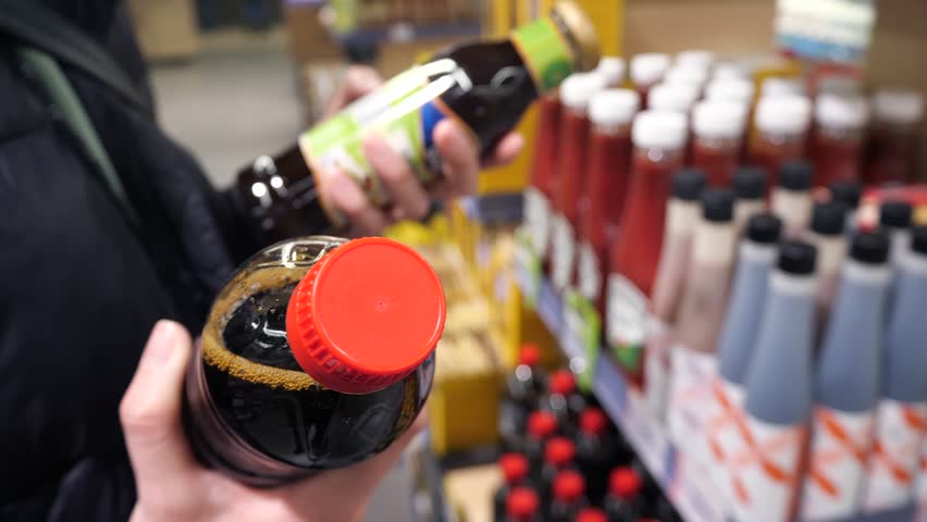 A young woman studies the labels of soy sauce bottles in a store and chooses one. Japanese cuisine, choosing a product in a store, comparison.