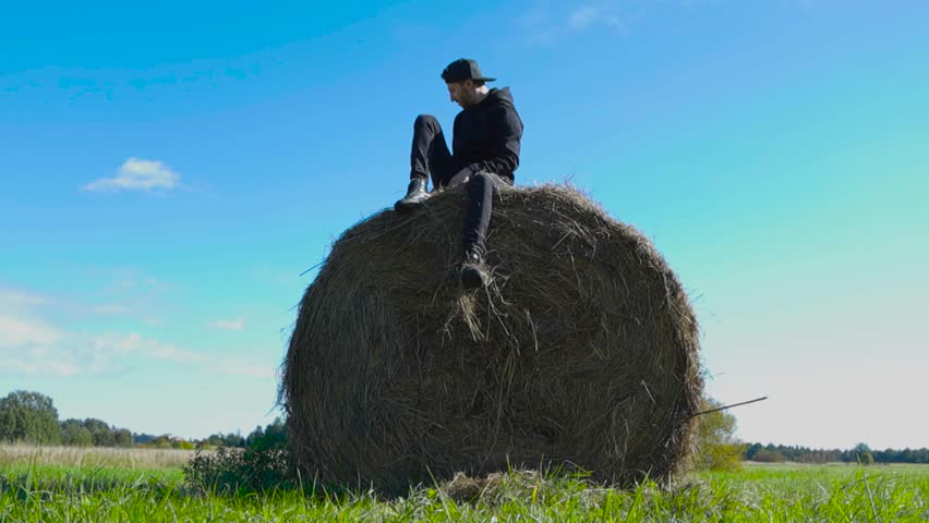 Caucasian man with black clothes sitting on top of a hay bale in a farm field and drops hay to the ground in slow motion from his hands. Summer time and sun is shining with blue sky in the background.
