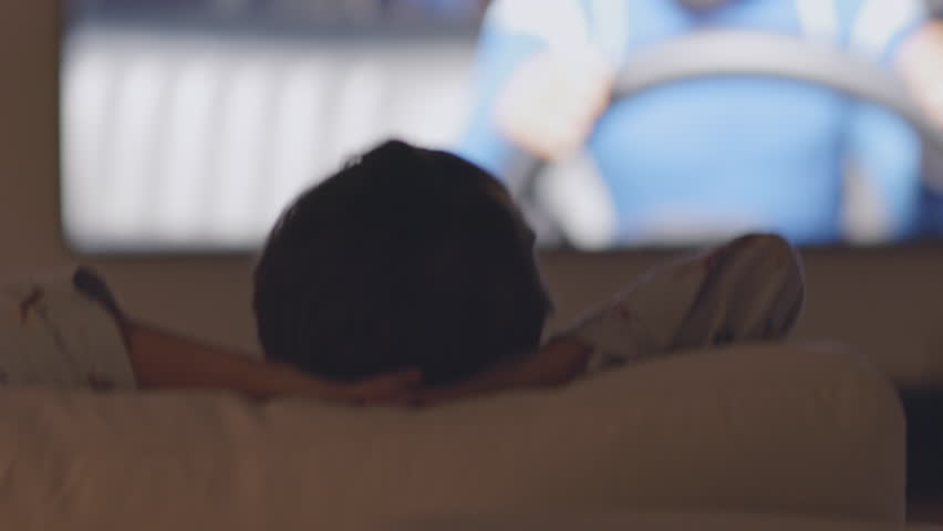 A young boy in pajamas relaxes on a couch, leaning back with hands behind his head, watching cartoons on a TV screen at night.
