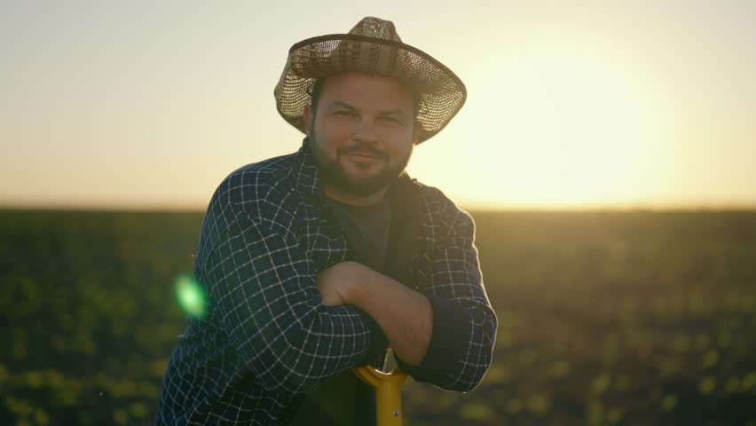 Joyful farm worker leaning on shovel after hard working in field, portrait. Smiling middle-aged man in straw hat looking at camera, successful farmer in beautiful agricultural field, eco agribusiness