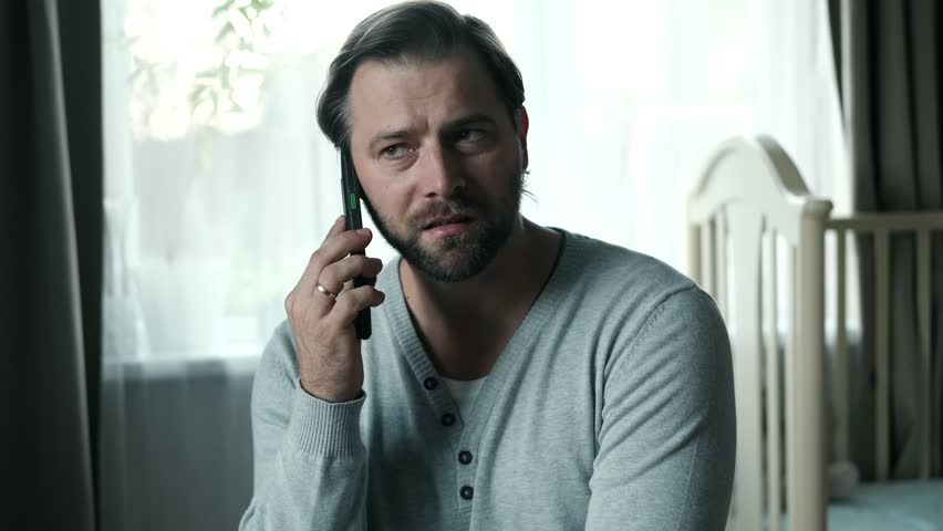 Emotional handsome bearded man talking on a mobile phone among a serene home environment. Thoughtful confident 30s male having conversation making call on smartphone. Men sitting on a bed with Cell.