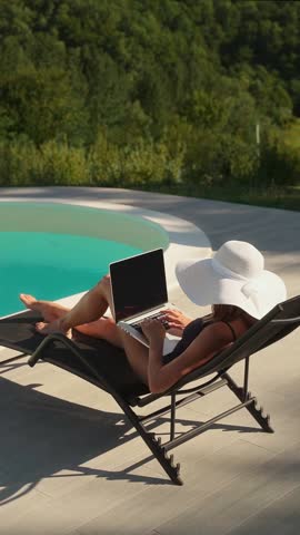 A young woman relaxes on a lounge chair near a pool on sunny day, using her laptop outdoors with a scenic green background. Work-life balance concept 