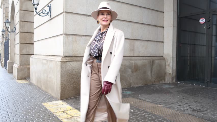 Stylish Mature Woman Wearing Fashionable Clothing Beige Coat and trousers, Felt Hat, Accessories Belt and Leather Gloves Walks Against Beige Wall in City, Street Style and Fashion Trends