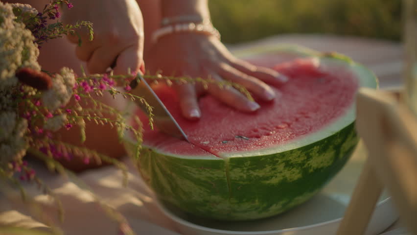Close-up of a hand firmly holding a juicy, red watermelon while slicing through its vibrant flesh, fresh, green rind contrasts with pink interior as individual cuts the fruit