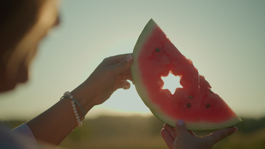 Close-up of woman holding watermelon slice with star cutout shape as sunlight shines through, creating a beautiful light effect and glowing silhouette