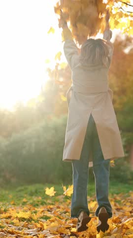 Carefree woman whirling in falling maple leaves in forest in sunny autumn day. Happiness and joy, good mood in fall season, young adult lady admiring beauty of nature, dry yellow foliage flying around
