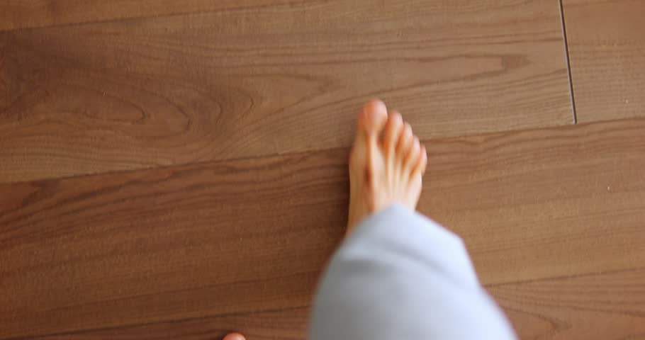 Man with bare feet walks on wooden floor of room with bright sun glare. Young guy in light grey trousers walks on parquet passing by chair slow motion