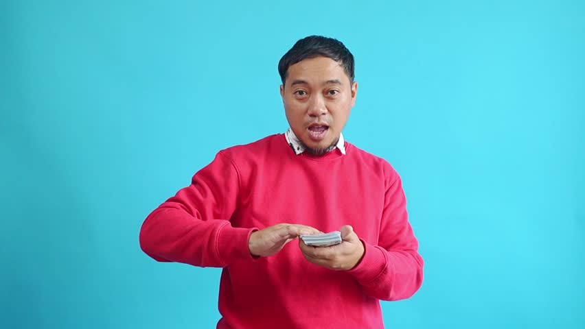 Happy Young Asian Man Throwing Money Banknotes On Camera Isolated On Light Blue Studio Background