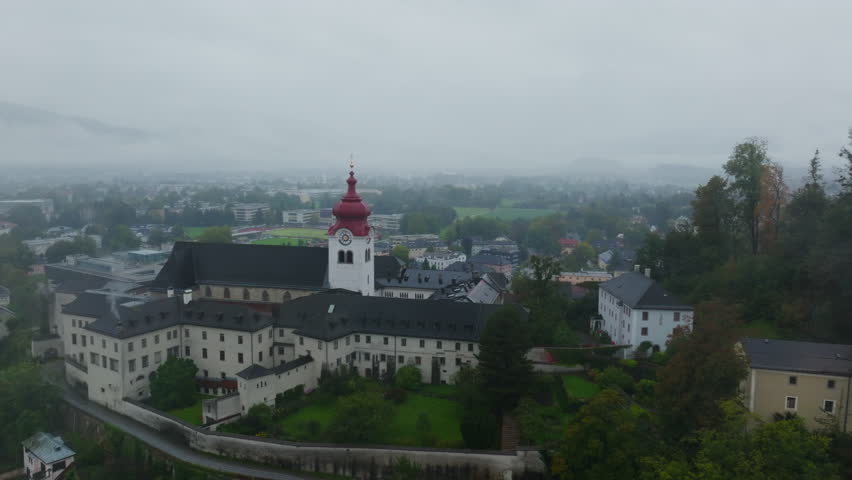 Aerial views of Salzburg, Austria showcase Nonnberg Abbey on a foggy and rainy day. Low clouds and mist create a mystical atmosphere over the historic architecture and scenic landscape