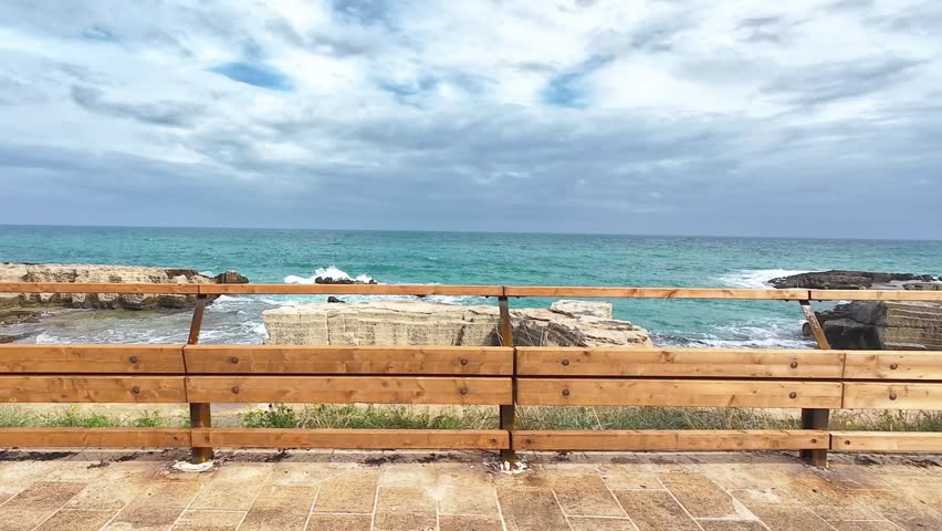 scenic drive along the coast with views of the blue ocean, a wooden fence, and greenery passing by. Gentle waves and a partly cloudy sky add to the serene seaside atmosphere