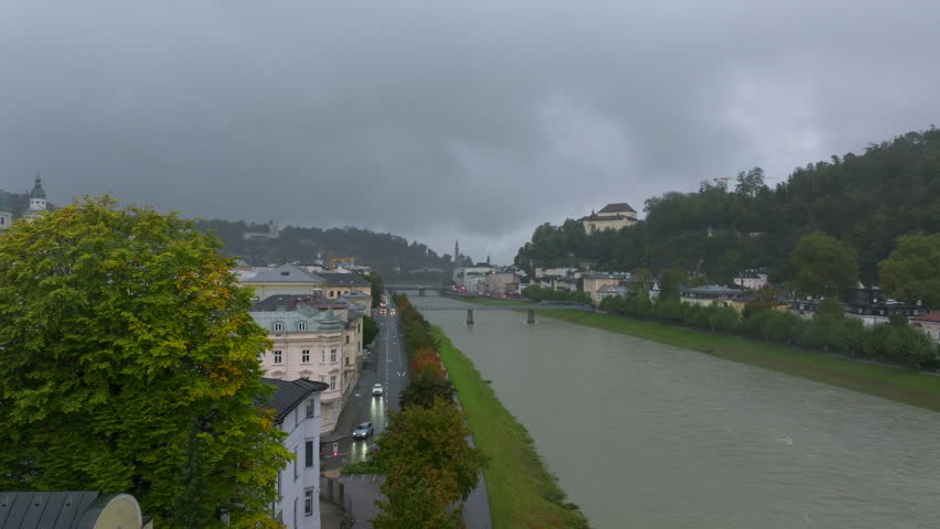 Aerial views showcase the Salzach River flowing through Salzburg, Austria, with buildings and a bridge lining the riverbank under a cloudy sky, creating a picturesque urban landscape