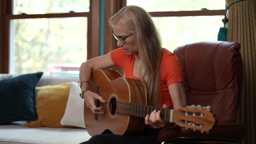 Mature woman with long blonde hair, wearing glasses and an orange shirt, sits in a brown leather armchair and plays an acoustic guitar. The room has a couch with pillows and a window