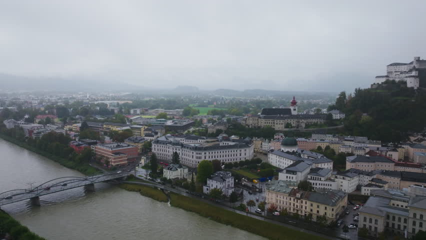 Aerial views of Salzburg, Austria showcase the Salzach River and Hohensalzburg Fortress under a cloudy sky, highlighting the city