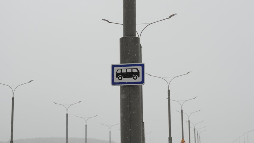 Bus stop sign on pole with falling snow, surrounded by numerous street lamps in muted, snowy landscape. Essence of winter in urban setting, highlighting impact of snow on public transportation.