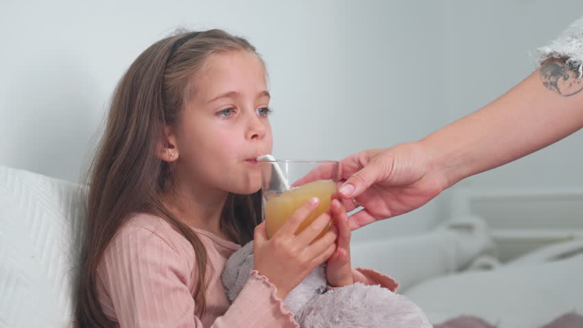 Sick girl drinking juice with straw from glass given by mother while sitting in bedroom at home. She is hydrating herself as she looks away while relaxing 