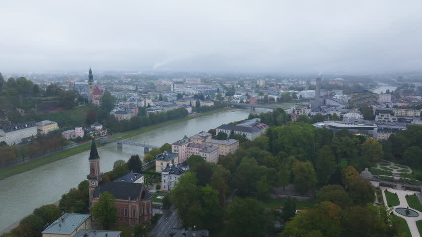 High angle tripod view of Salzburg featuring the Salzach River winding through the cityscape. Buildings and green spaces are visible under a cloudy sky